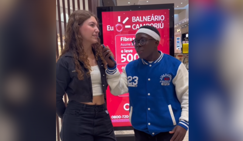 Jovem angolano vira cupido em Florianópolis - menino baixo, negro, usando casaco azul e branco entrevista menina branca, magra, que usa roupa preta e camisa cropped branca