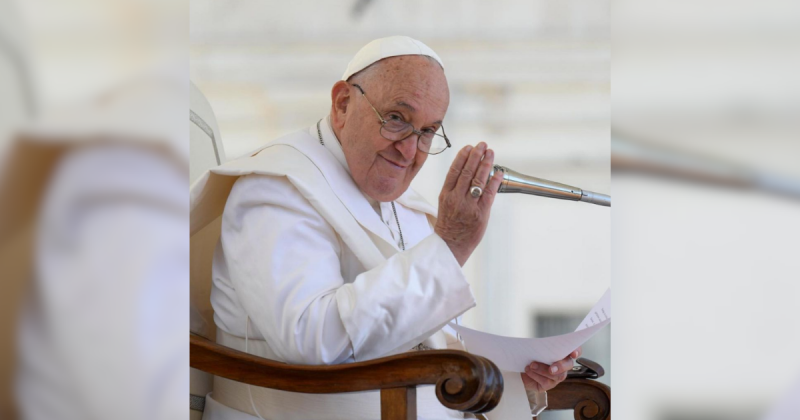 Papa Francisco acenando sentado em sua cadeira de madeira com microfone perto do rosto