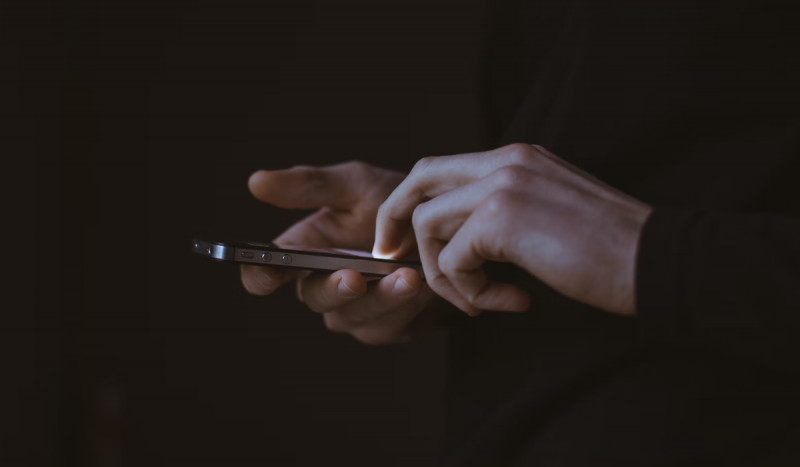 Mãos de pessoa branca mexendo em celular em frente a fundo preto