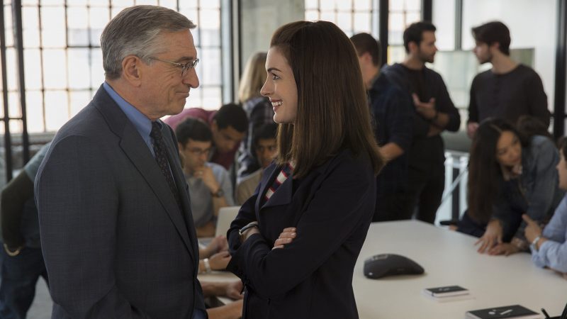 Imagem do filme 'Um senhor estagiário' em que há os personagens de Robert De Niro e Anne Hathaway virados um para o outro no escritório com pessoas trabalhando ao fundo.