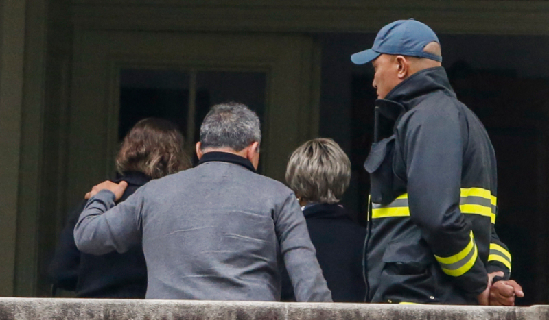 Familiares de vítimas do acidente com avião da Voepass - foto mostra homem de casaco cinza de costas segurando no ombro de mulher também de costas usando camisa preta, enquanto um homem com casaco preto e listra amarela no meio olha eles saindo