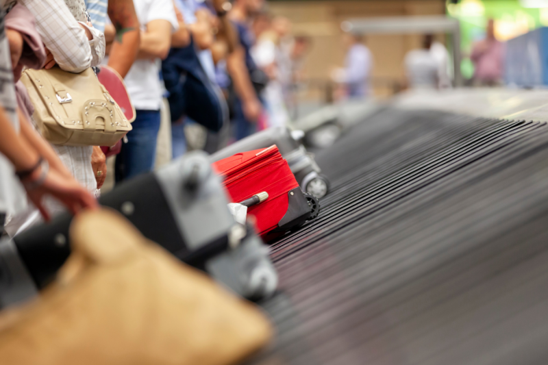 A foto mostra parte da esteira de um aeroporto com malas em cima. O foco está sobre uma mala vermelha. As demais aparecem desfocadas. Ao lado, há uma fila de pessoas. Não aparece o rosto delas, apenas braços, tronco e pernas. 